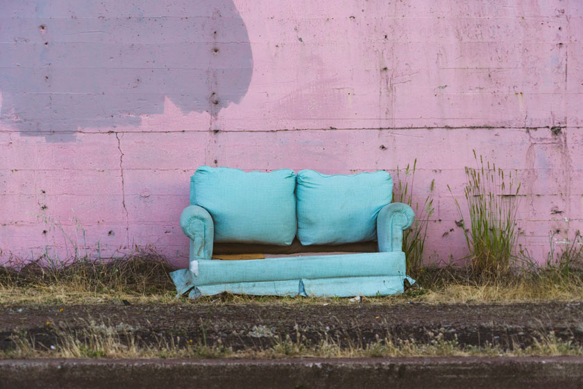 Blue sofa couch with no bottom cushions sitting against pink building in grass.