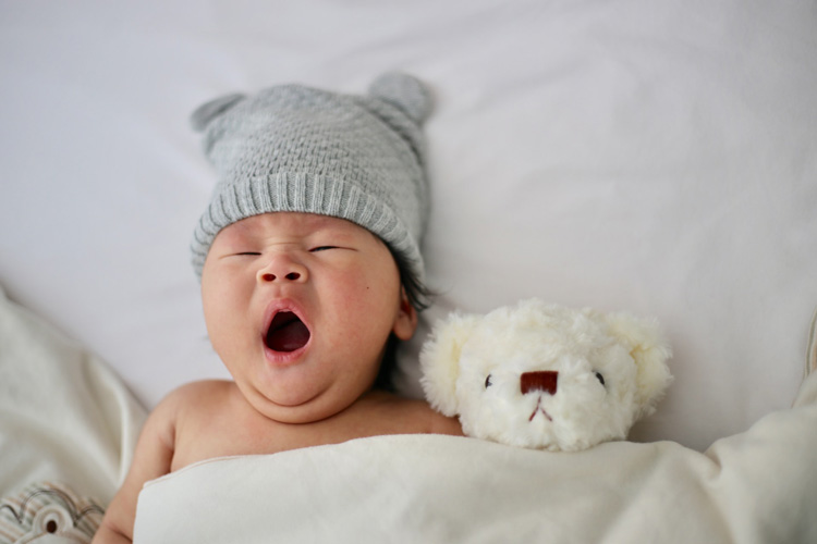 A baby wearing a knit hat tucked in bed with a teddy bear